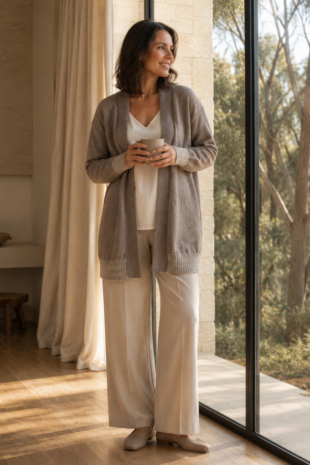 A woman stands by a large window in a bright room, wearing a light taupe knit cardigan over a white top and cream wide-leg pants. She holds a white mug, with warm sunlight streaming in from the right side.