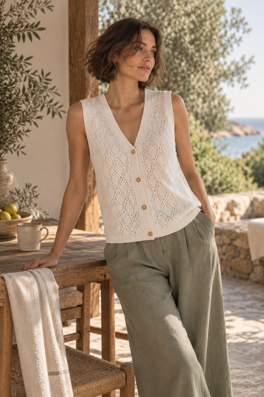 A woman stands on a sunlit outdoor patio wearing a sleeveless cream knit cardigan with gold buttons and olive green wide-leg pants.