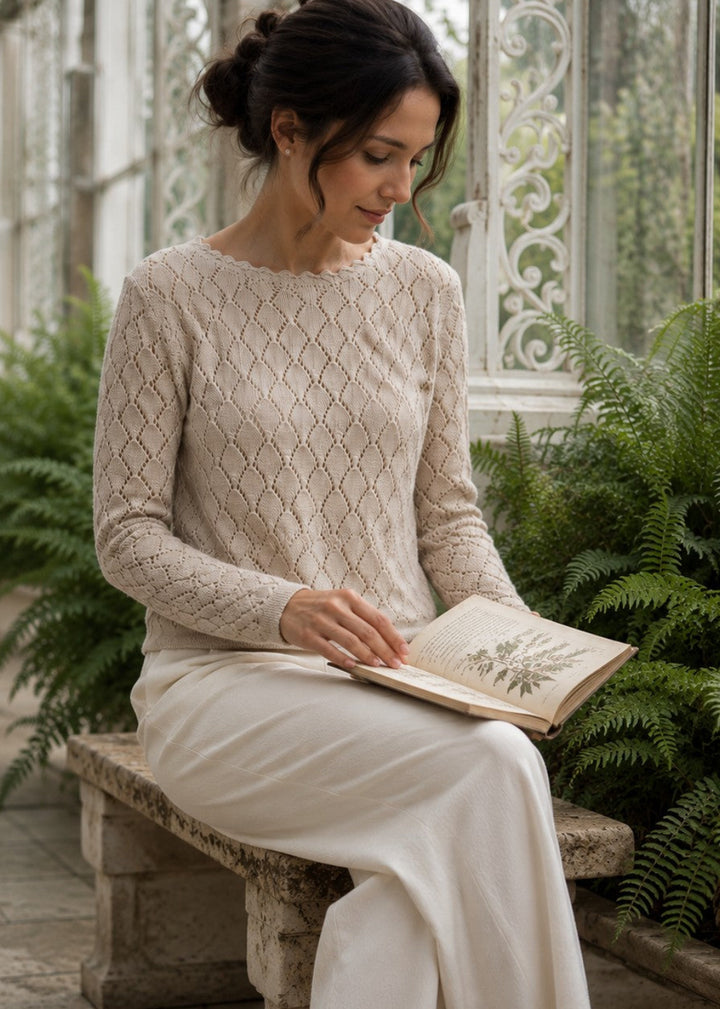A woman sits on a stone bench in a greenhouse, wearing a cream cable-knit long-sleeve sweater and matching cream wide-leg trousers while reading an open book surrounded by green ferns.