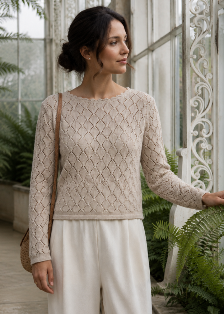 A woman stands in a greenhouse wearing a beige textured knit long-sleeve top and white wide-leg trousers, with a brown shoulder bag.