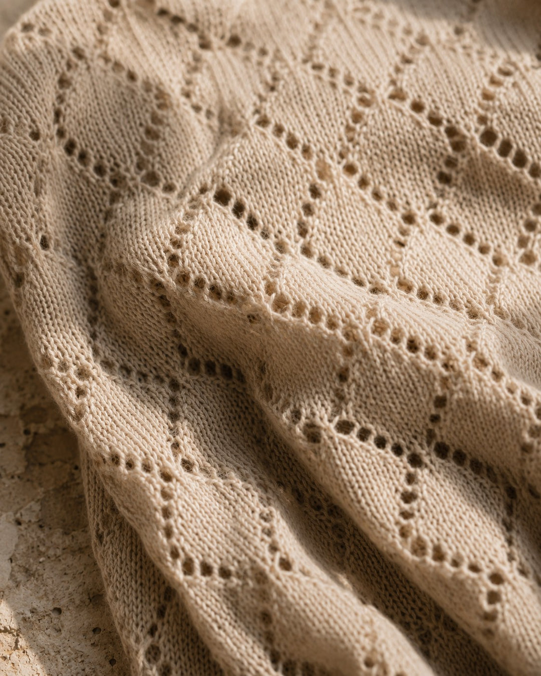 Close-up of a beige knit fabric with an openwork diamond lattice pattern, draped with soft folds and shadows.