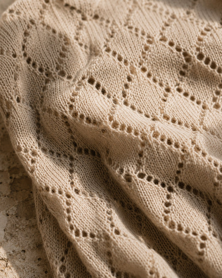 Close-up of a beige knit fabric with an openwork diamond lattice pattern, draped with soft folds and shadows.