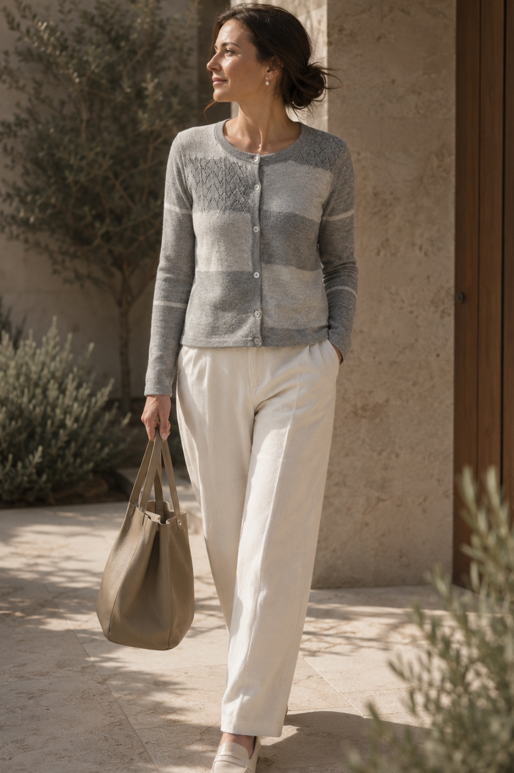 A woman walking outdoors in a sunlit courtyard wearing a gray and beige striped knit cardigan, cream trousers, and beige shoes, while carrying a taupe tote bag.