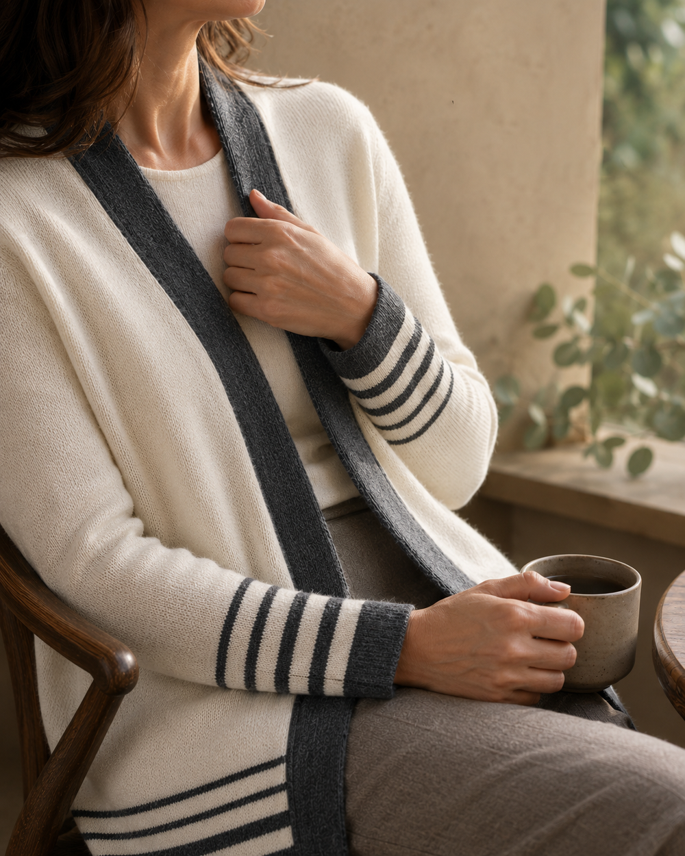 A woman seated indoors wearing a cream knit cardigan with dark gray striped trim and a dark gray knit scarf, holding a ceramic mug near a window.