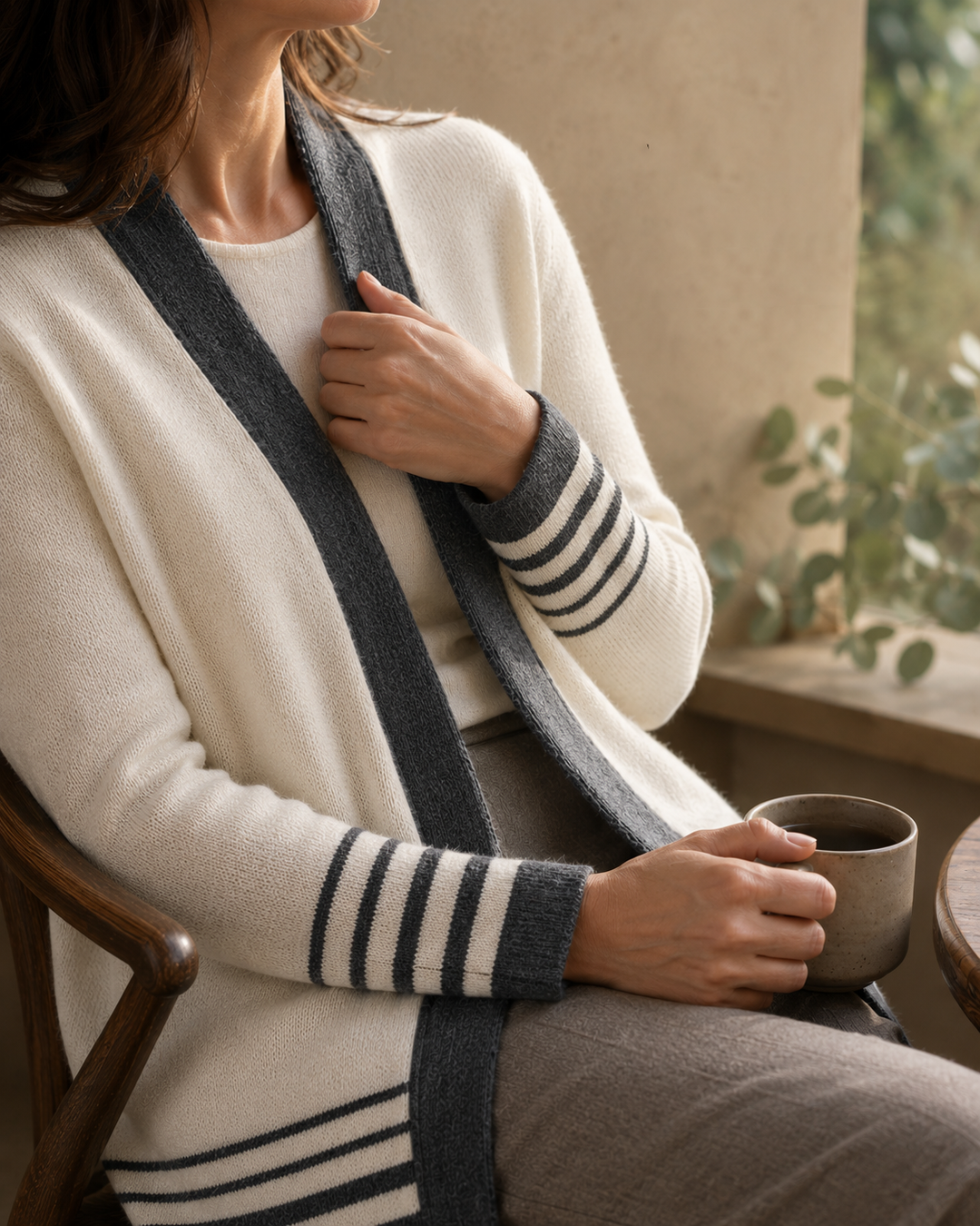 A woman seated indoors wearing a cream knit cardigan with dark gray striped trim and a dark gray knit scarf, holding a ceramic mug near a window.