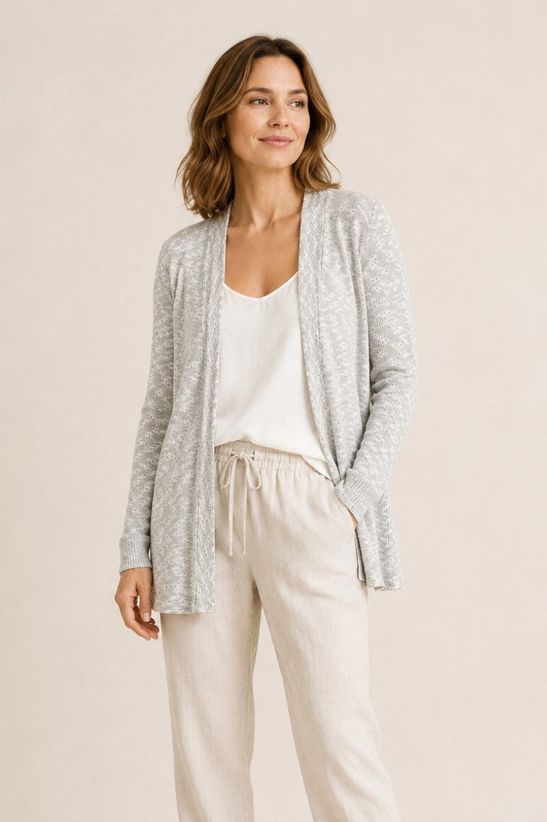 A woman modeling a light gray open-front cardigan over a white tank top and cream drawstring pants, paired with white sneakers, against a solid beige studio background.