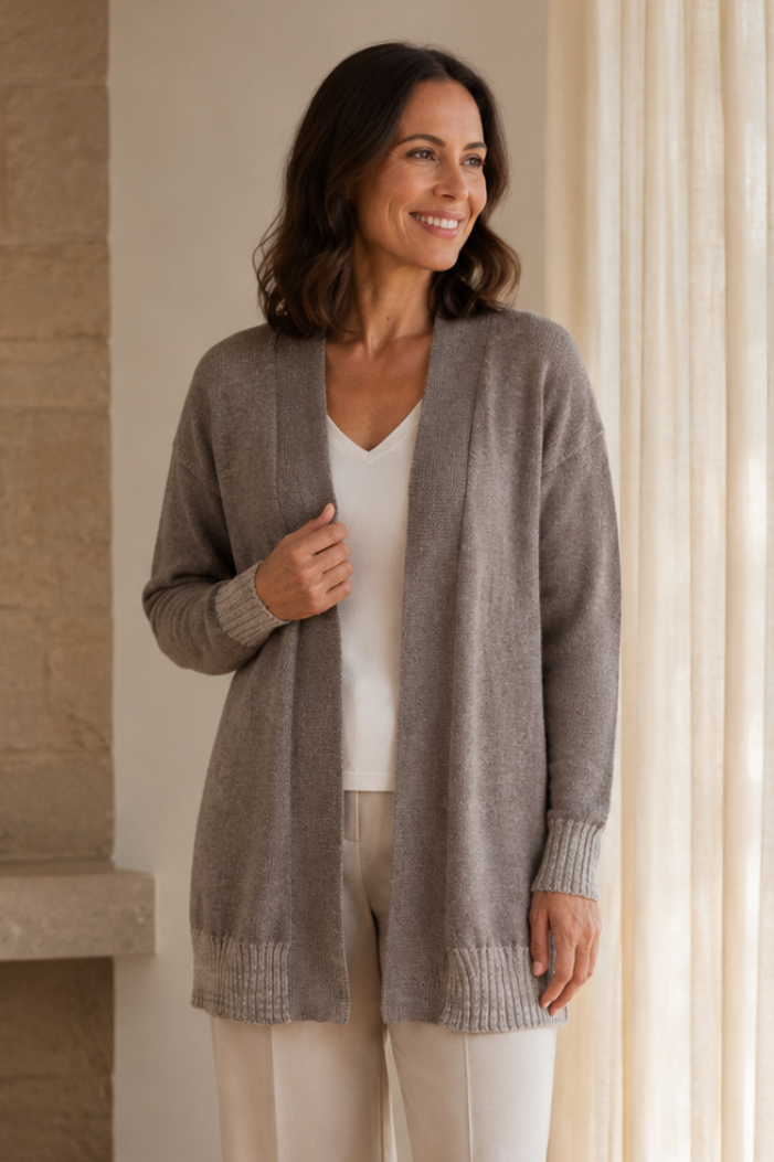 A woman stands in a bright room wearing a long taupe-gray knit cardigan over a white top and cream wide-leg pants.