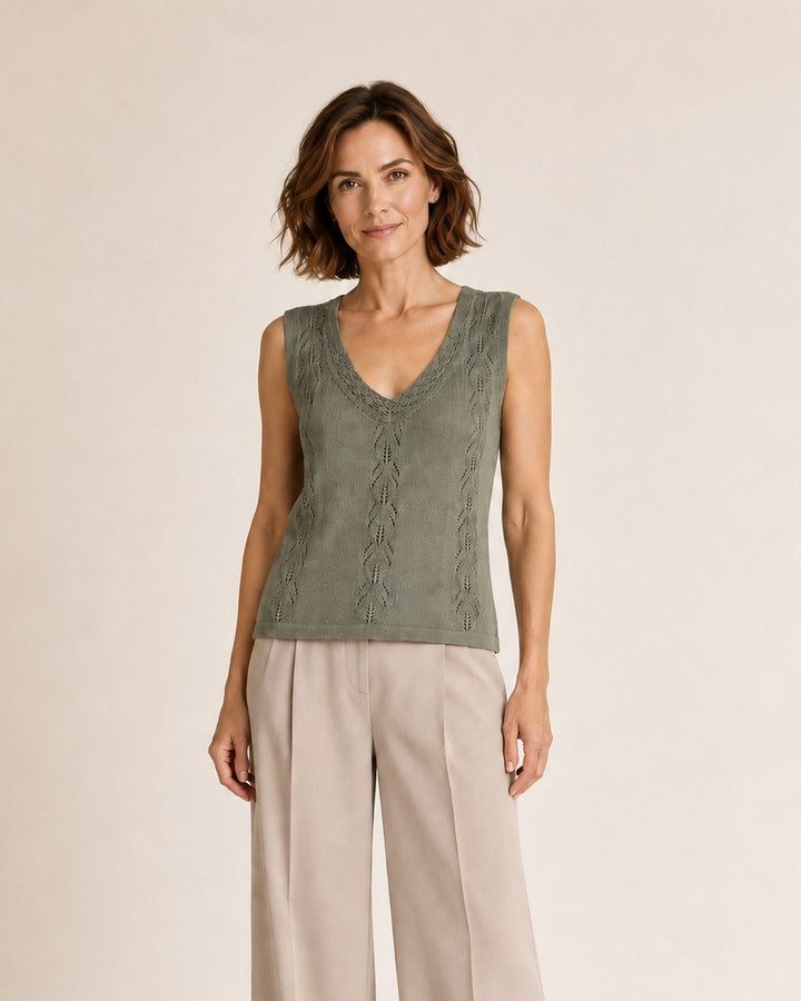 A woman modeling a sleeveless sage green knit vest with a V-neck and textured pattern, paired with wide-leg beige trousers, against a solid light beige studio background.