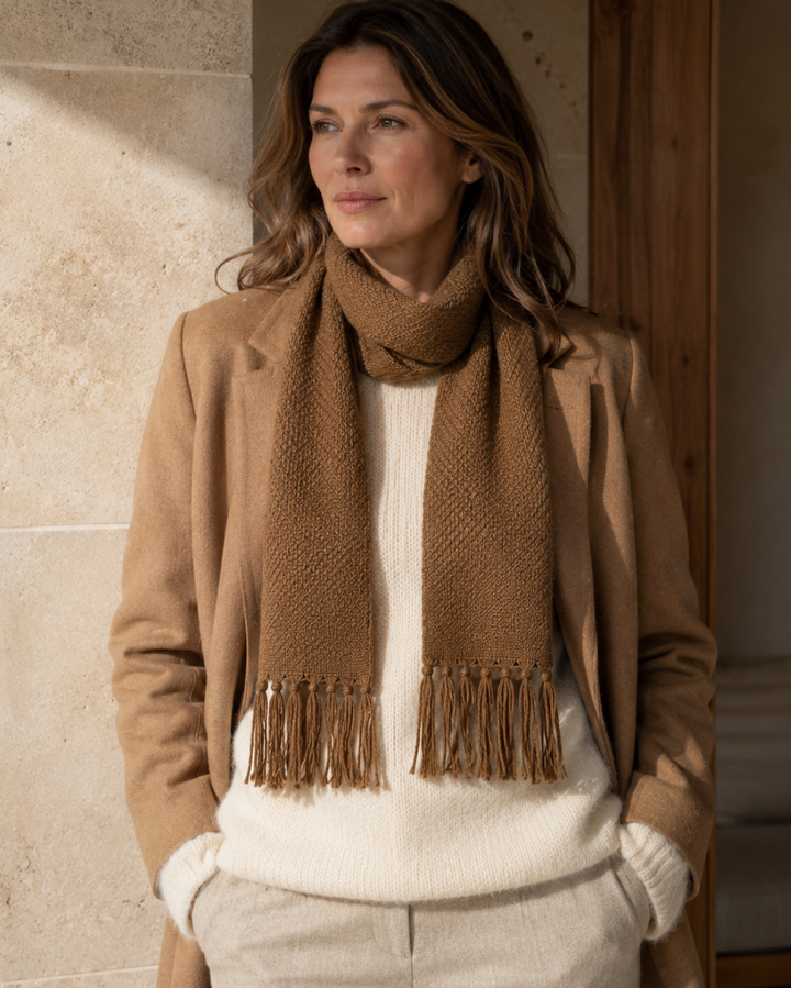 A woman wearing a camel brown knit scarf with fringe, styled over a cream ribbed sweater and beige trousers, standing in a warmly lit indoor setting.