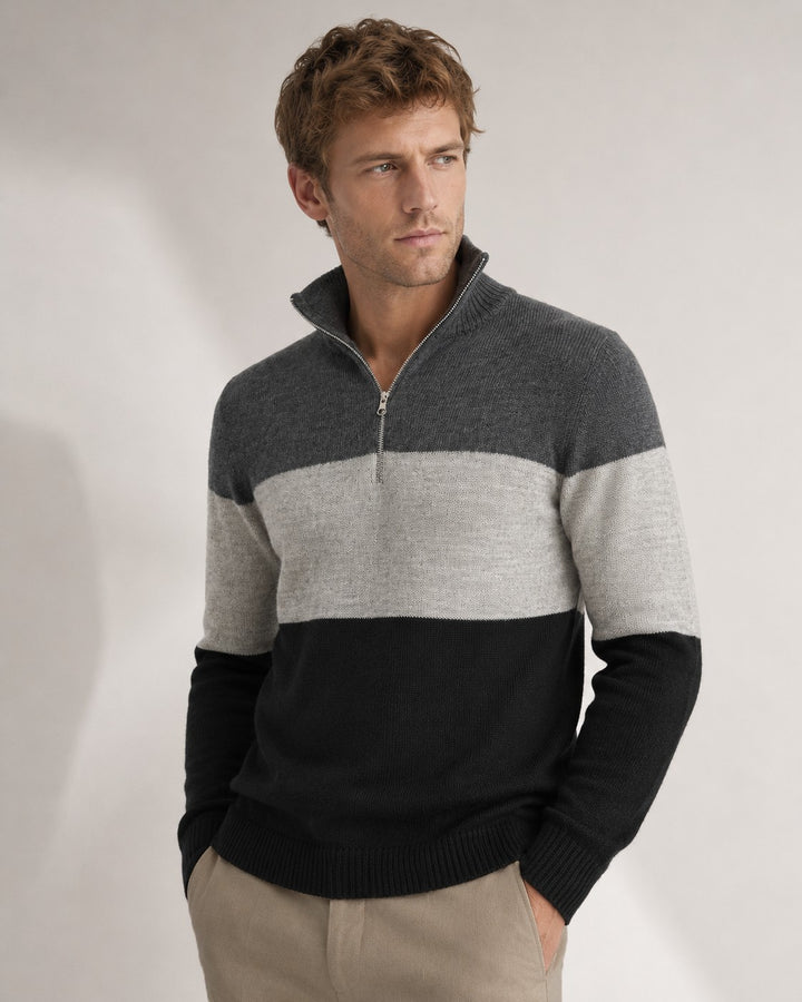 A man wearing a color-blocked quarter-zip knit sweater with dark gray, light gray, and black horizontal stripes, paired with beige trousers, against a neutral studio background.