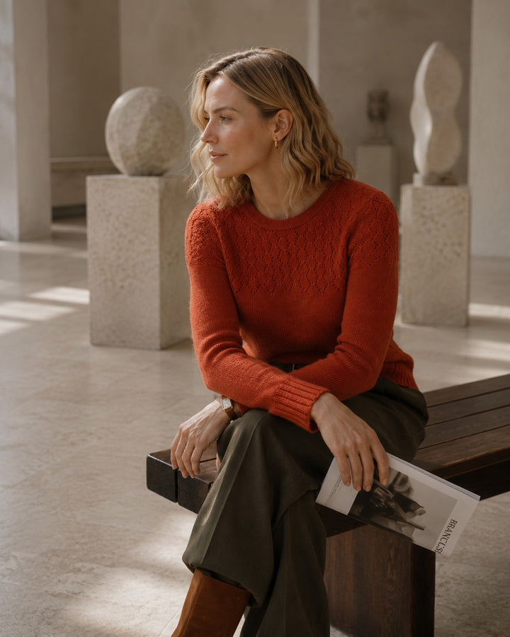 A woman sits on a wooden bench in a minimalist gallery, wearing a textured rust-orange crewneck sweater and dark olive green trousers, holding a magazine.