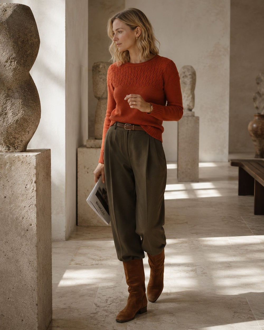 A woman stands in a sunlit indoor gallery wearing a textured rust-orange knit sweater, olive green wide-leg cropped trousers, and brown knee-high boots, holding a magazine.